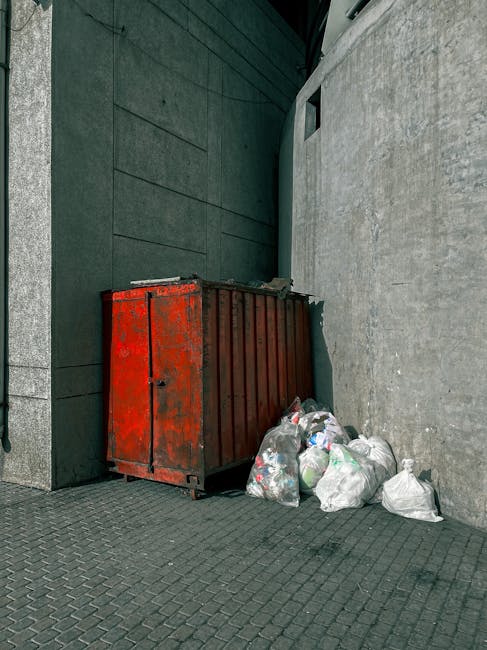 A sanitation worker wearing a high-visibility orange vest and dark trousers operates a large red rubbish collection truck, which is positioned on the side of a street. The truck's rear loading mechani