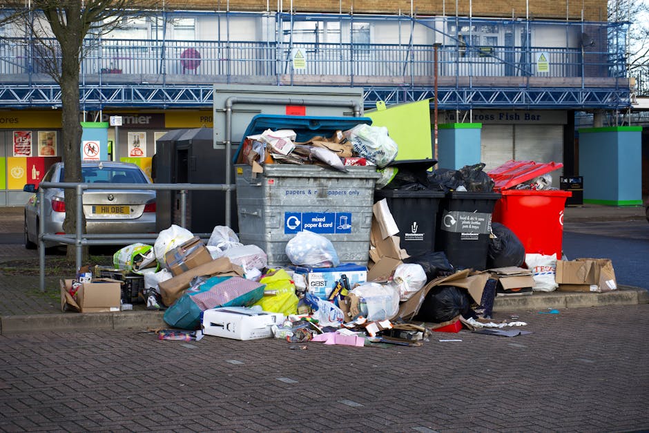 A collection of black rubbish bags, some partially torn, are piled against the base of a red metal door set into a plain, off-white textured wall. The bags appear to contain household waste or discard